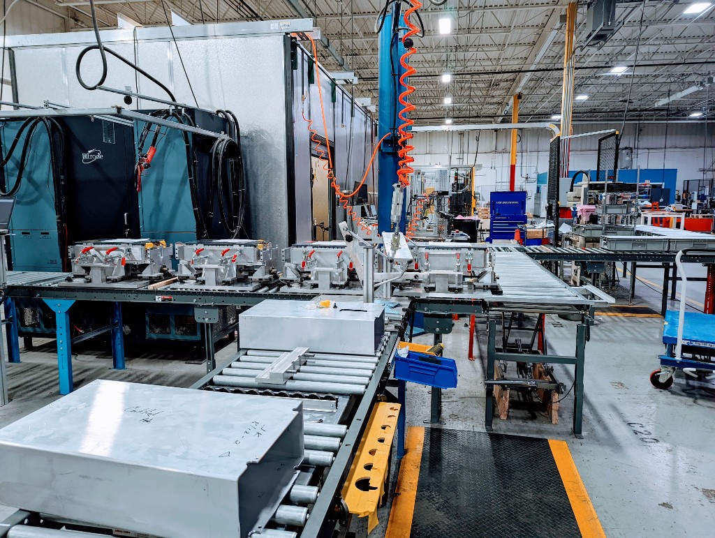 Battery pack assemblies on a roller conveyor in a high-bay assembly and validation area, with industrial charging equipment, environmental chamber, overhead utilities, and safety-orange accents on the floor and services.